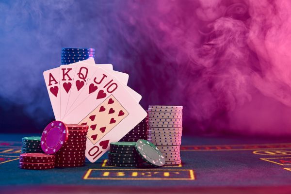 Winning combination in poker, hearts, standing leaning on multicolored chips piles on a blue cover of a playing table with markup. Black, smoke background with red and blue backlights. Gambling entertainment, playing cards, casino concept. Close-up shot.